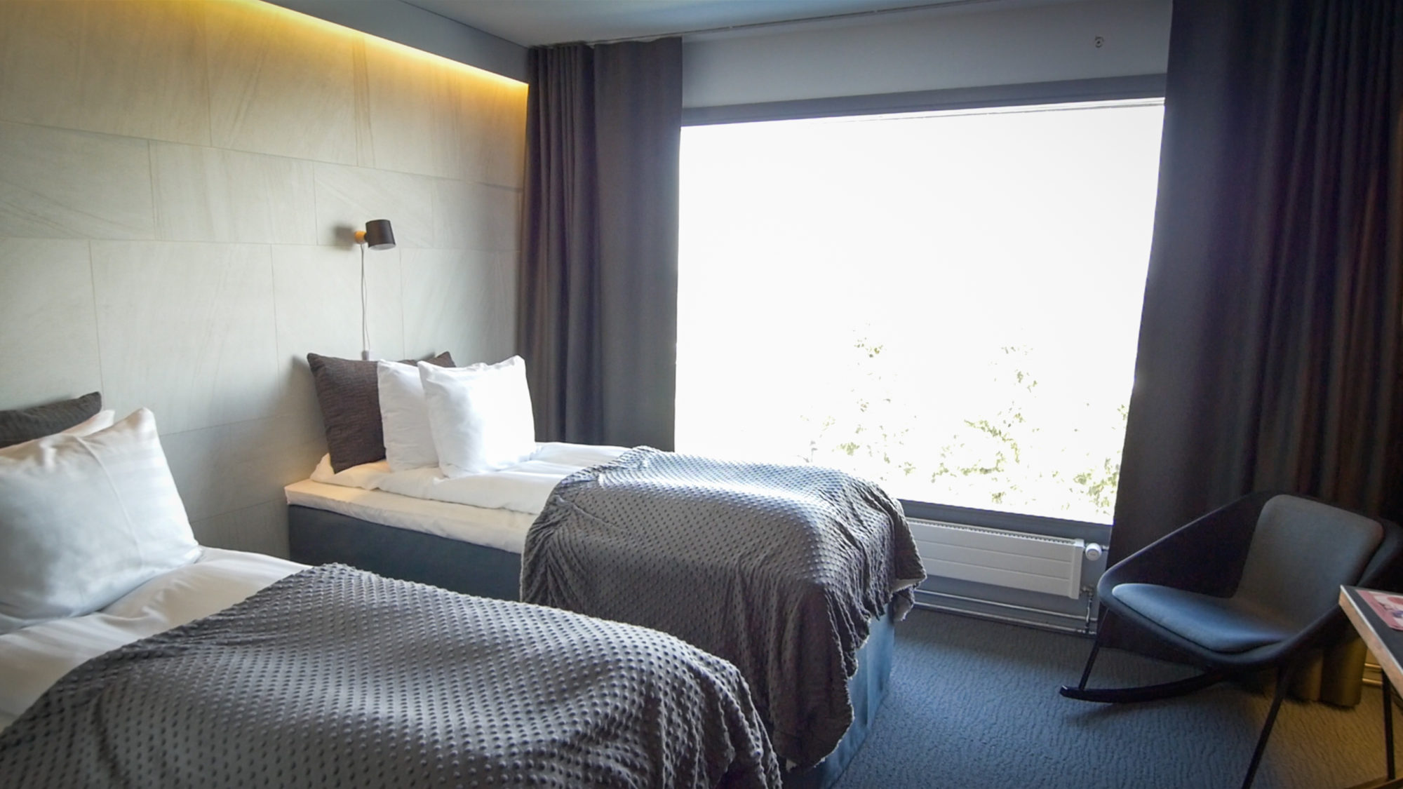 Remontoitu hotellihuone jossa vaalea laattaseinä ja tummanharmaa, aaltokuvioitu kokolattiamatto.